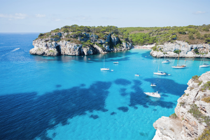 Barcos y veleros fondeados en una cala de aguas turquesas rodeada de acantilados